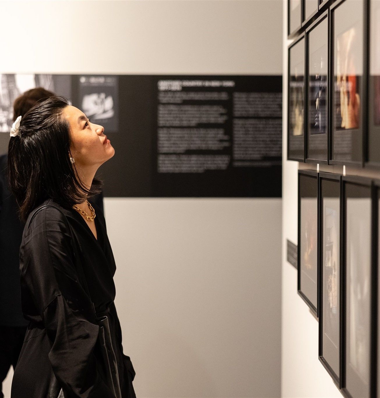 a woman with black hair looks up towards a wall with pictures in black frames