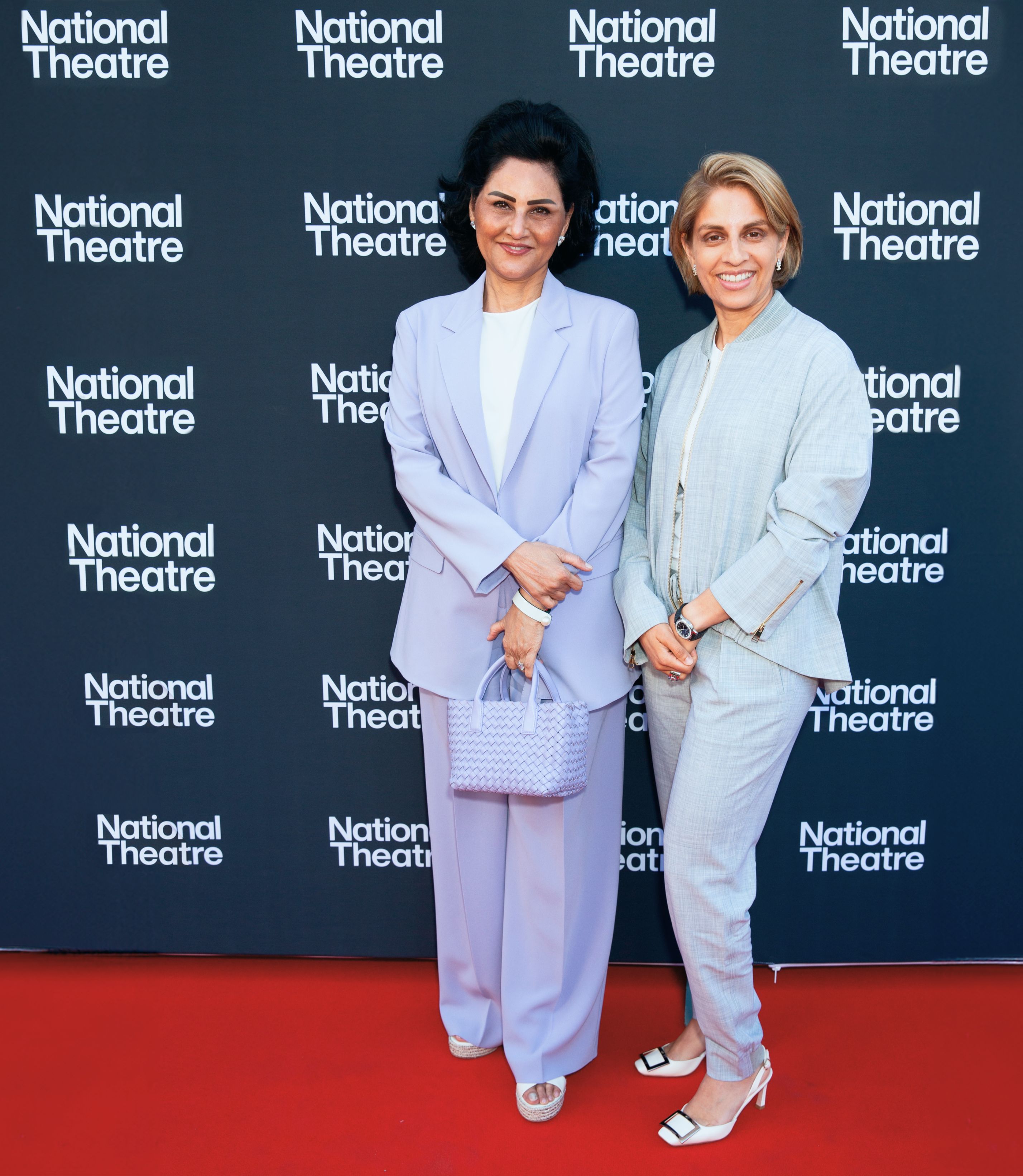Two women of South Asian ethnicity stand on a red carpet. One has black, shoulder length hair and wears a lilac suit. The other has a blonde bob and wears a light gray suit.